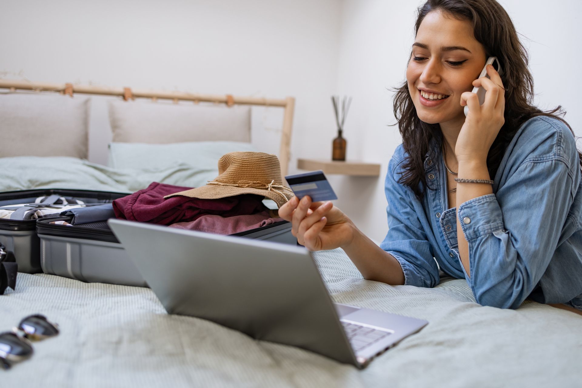 Woman on phone, holding a credit card, looking at laptop while packing suitcase on bed.