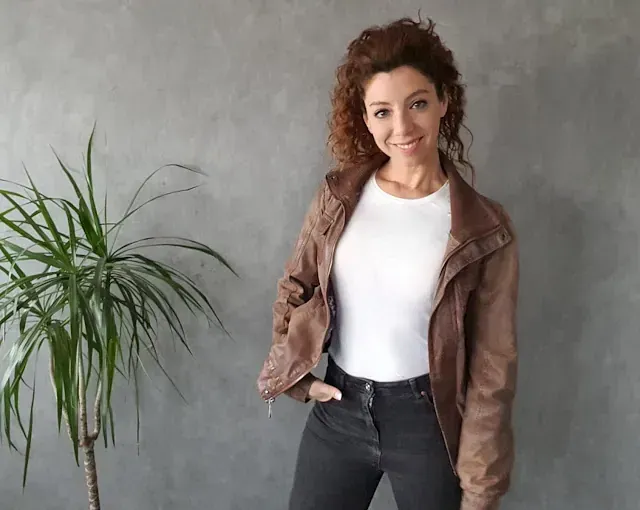 Una mujer que viste una camisa blanca y una chaqueta marrón está parada frente a una planta.
