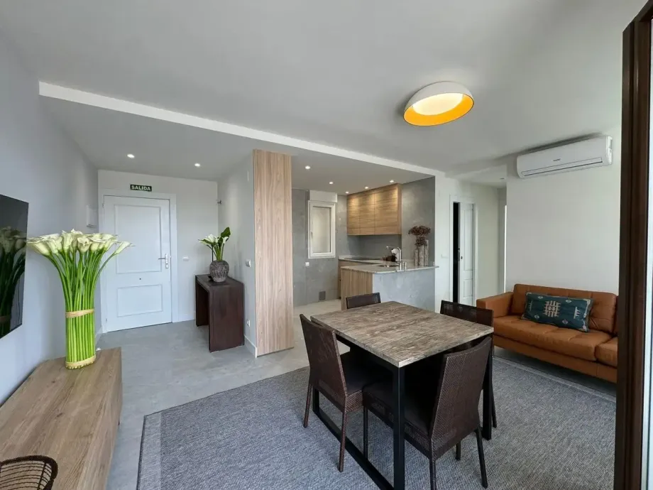 Acogedor espacio de estar: comedor con mesa, cocina al fondo, sofá de cuero, flores, tonos neutros.