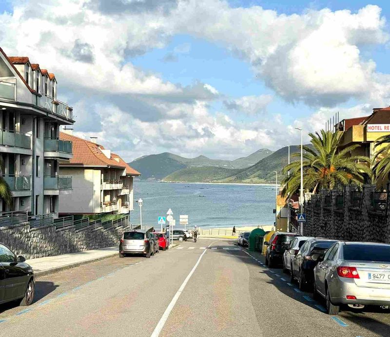 La calle conduce a la playa con coches aparcados, edificios y montañas bajo un cielo nublado.