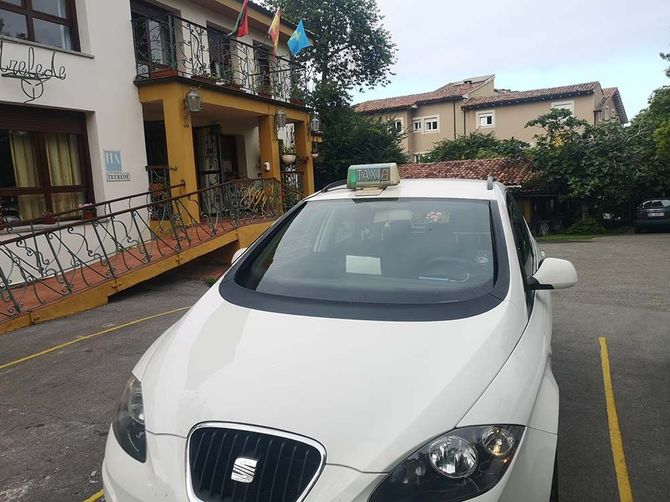 Taxi blanco estacionado frente a un edificio amarillo con rampa y banderas ondeando encima.