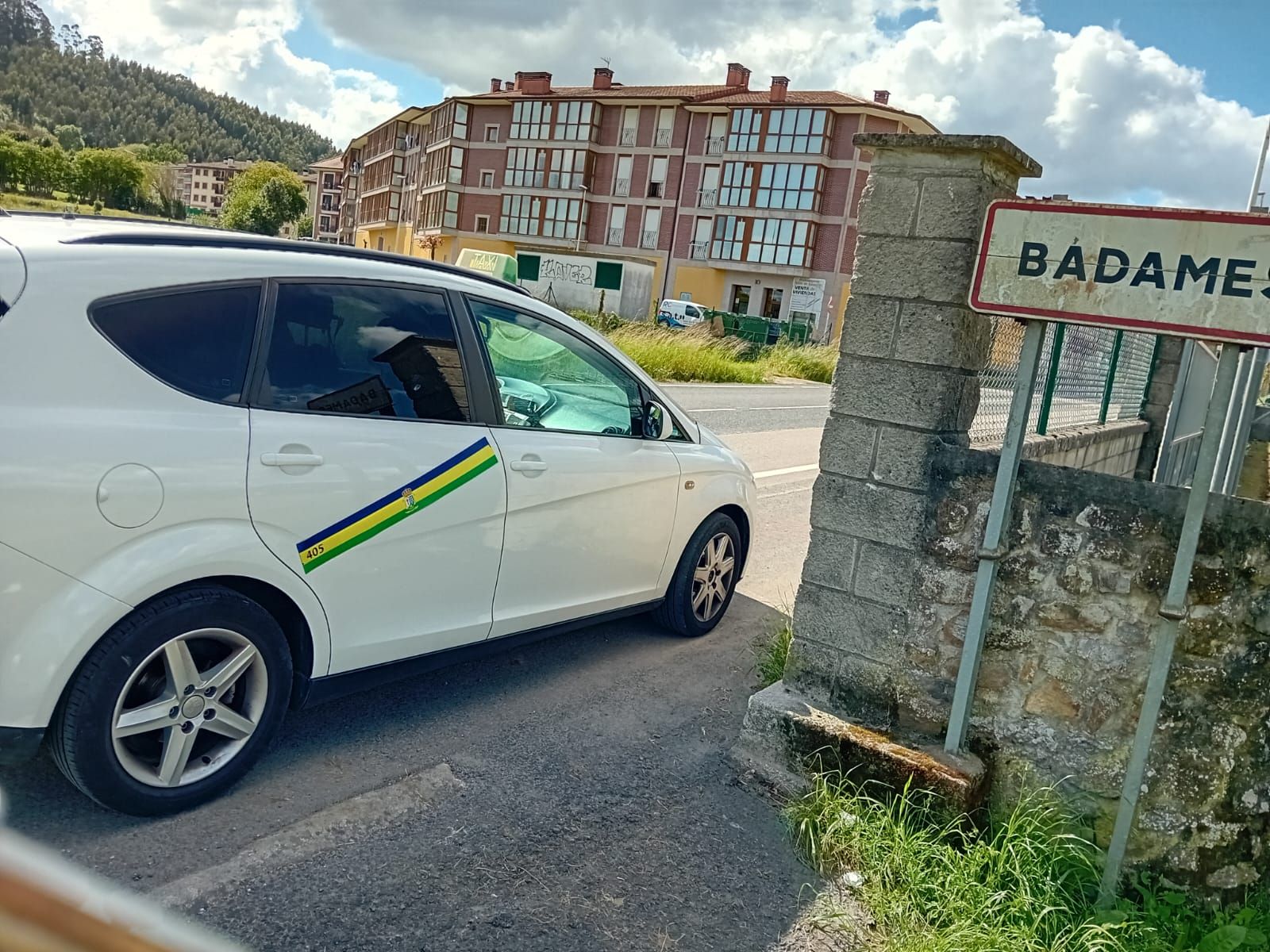 Coche blanco con franja verde y amarilla junto a un cartel de Badame. Edificio al fondo.