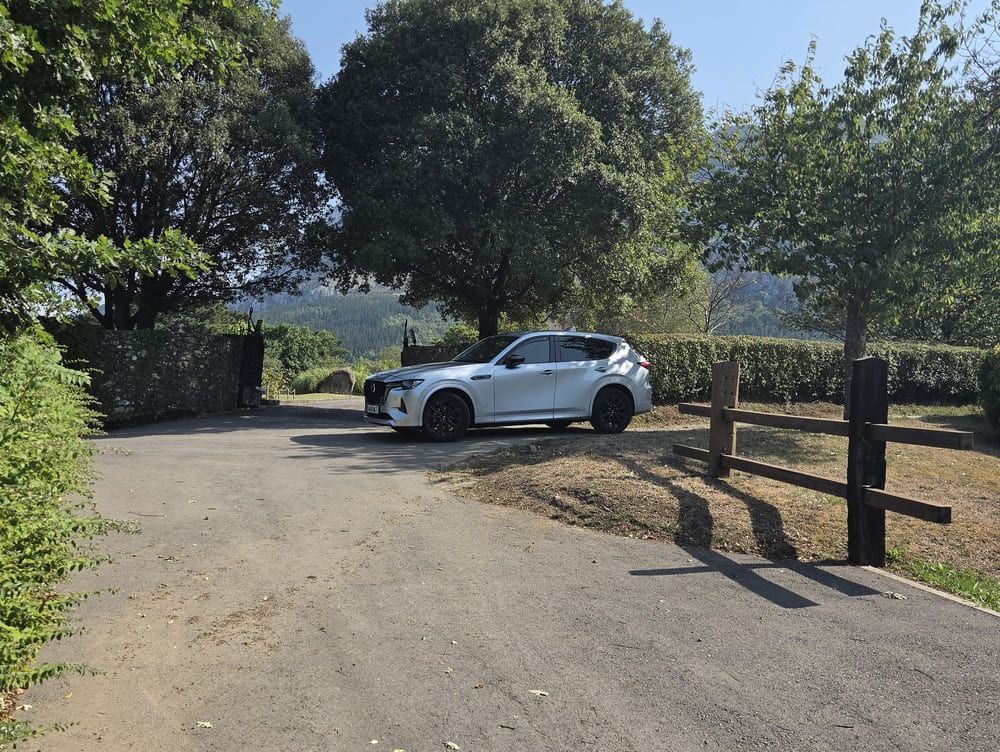 Un SUV blanco estacionado en un camino de tierra soleado junto a árboles y una cerca de madera, con colinas en la distancia.