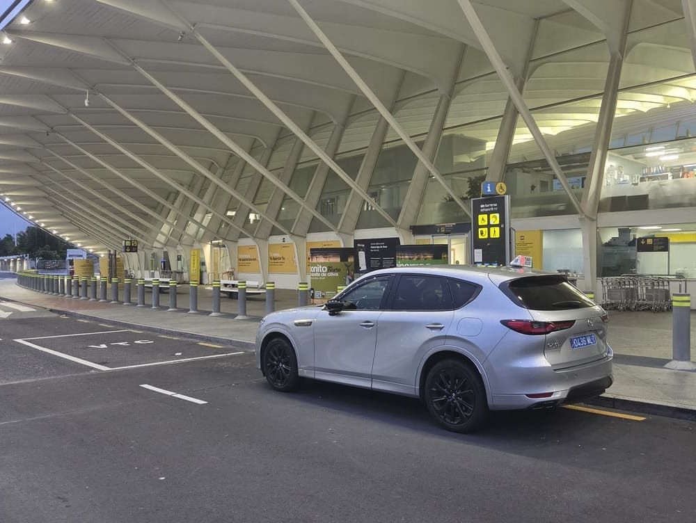 SUV plateado en una terminal de aeropuerto junto a la acera, con carriles de facturación y una pasarela cubierta al fondo.