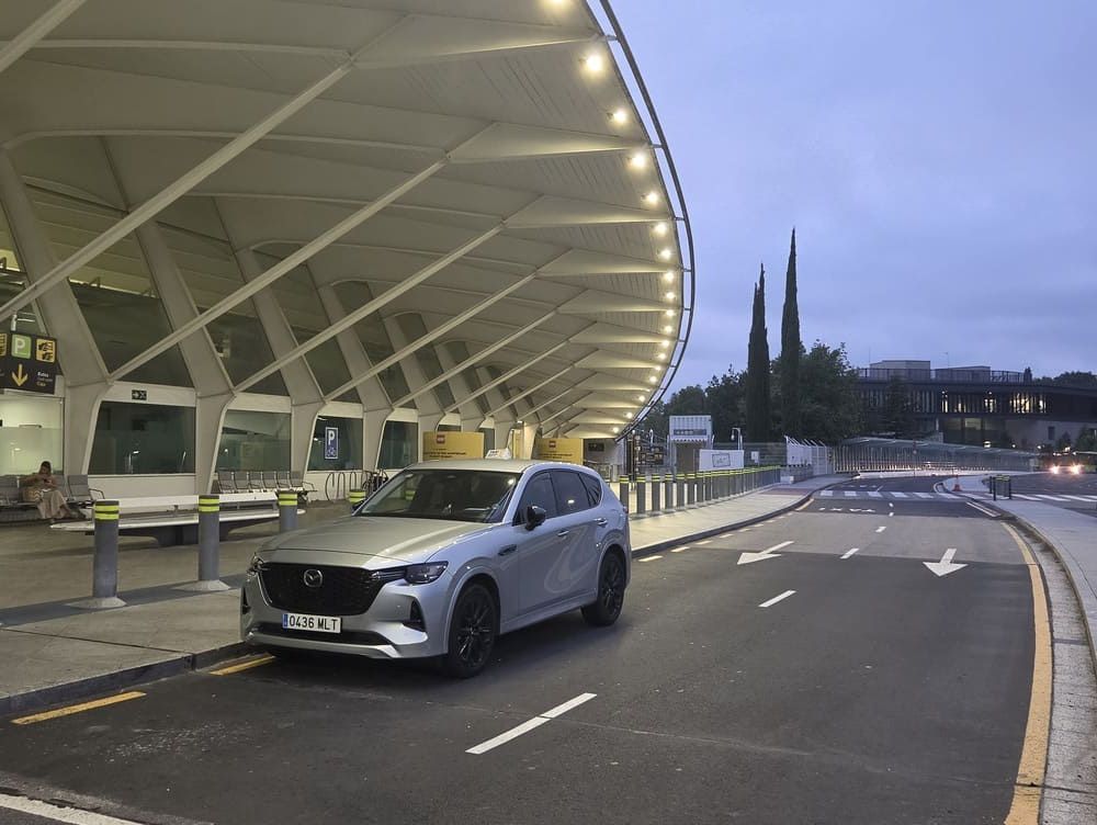 Un SUV plateado estacionado en la acera de un aeropuerto moderno al anochecer, bajo una marquesina iluminada.