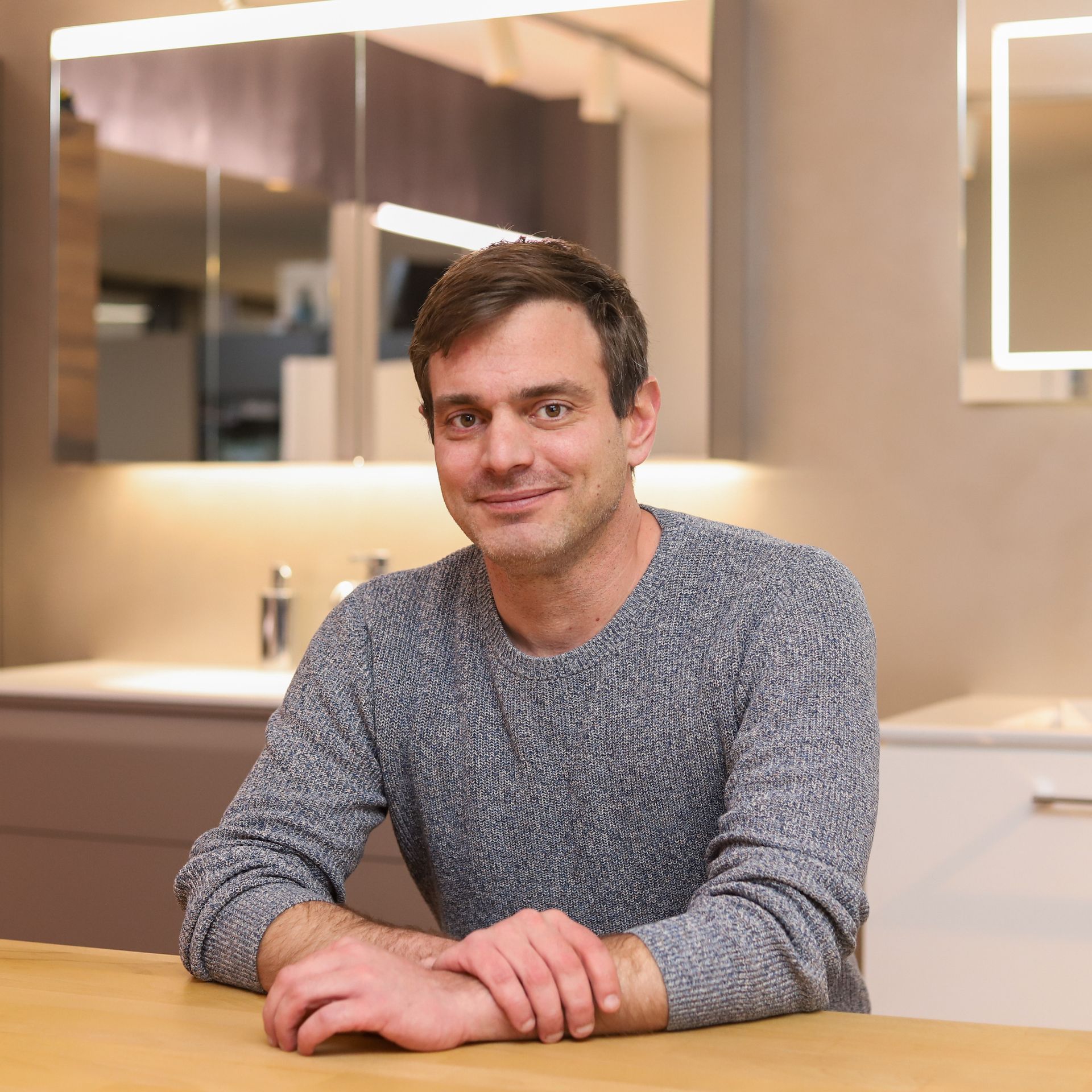 Ein Mann in einem grauen Pullover sitzt mit gefalteten Händen an einem Tisch