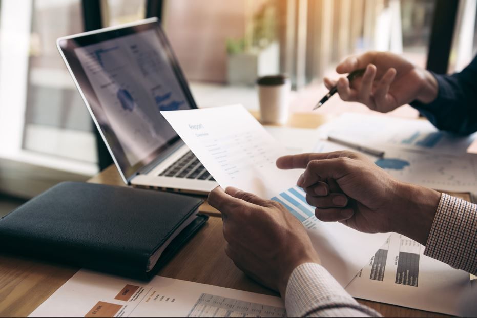 Dos profesionales analizan gráficos y documentos de datos en un escritorio con una computadora portátil y un cuaderno.