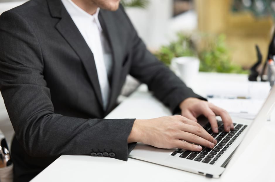Una persona vestida con un traje oscuro teclea en un ordenador portátil en un escritorio blanco en una oficina luminosa.