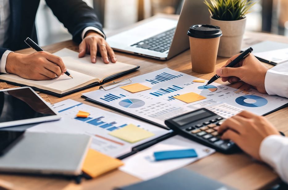 Personas trabajando juntas en un escritorio con computadoras portátiles, calculadoras y gráficos financieros.