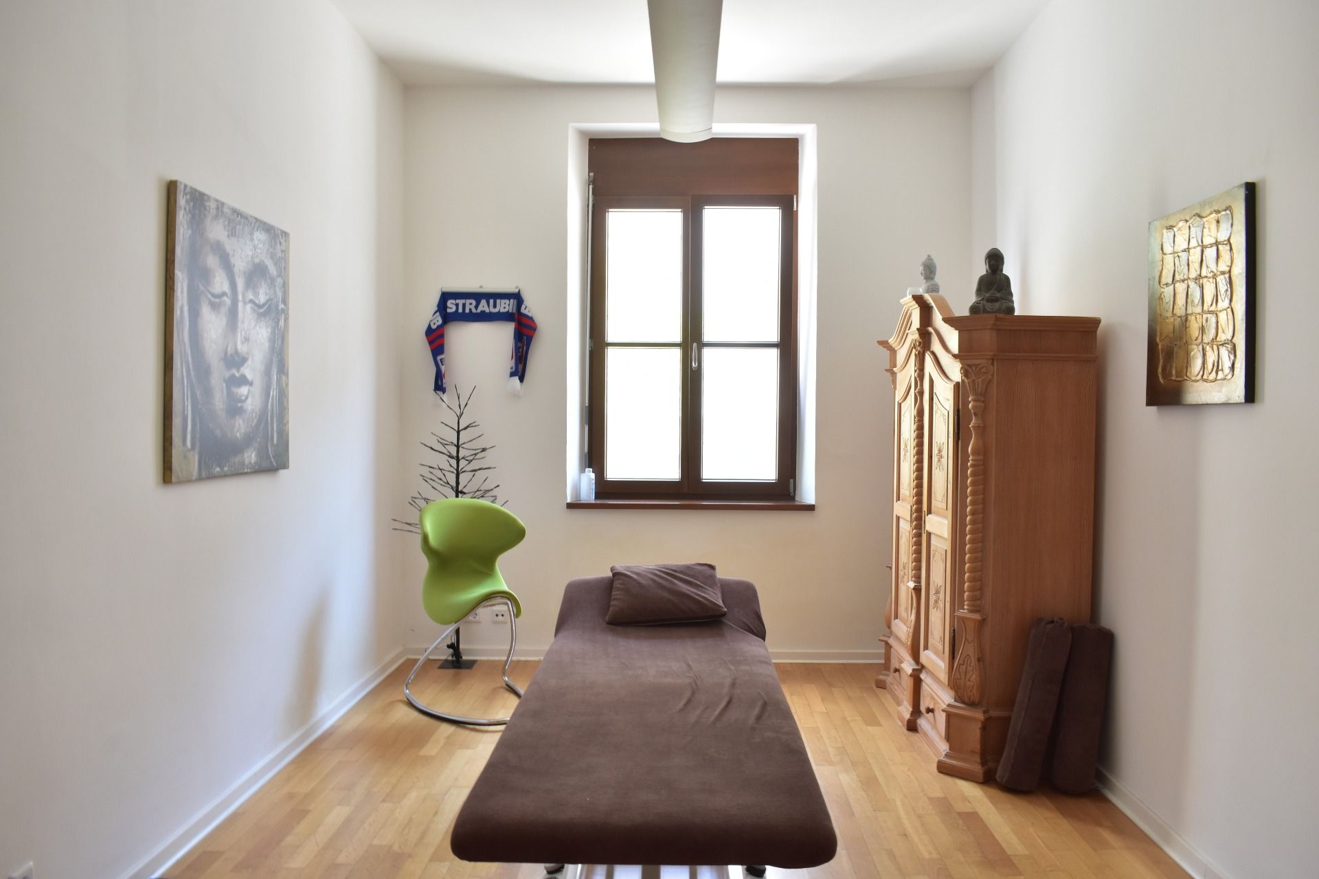 Massageraum: Massageliege, Fenster, Kunstwerk, Holzschrank, grüner Stuhl.