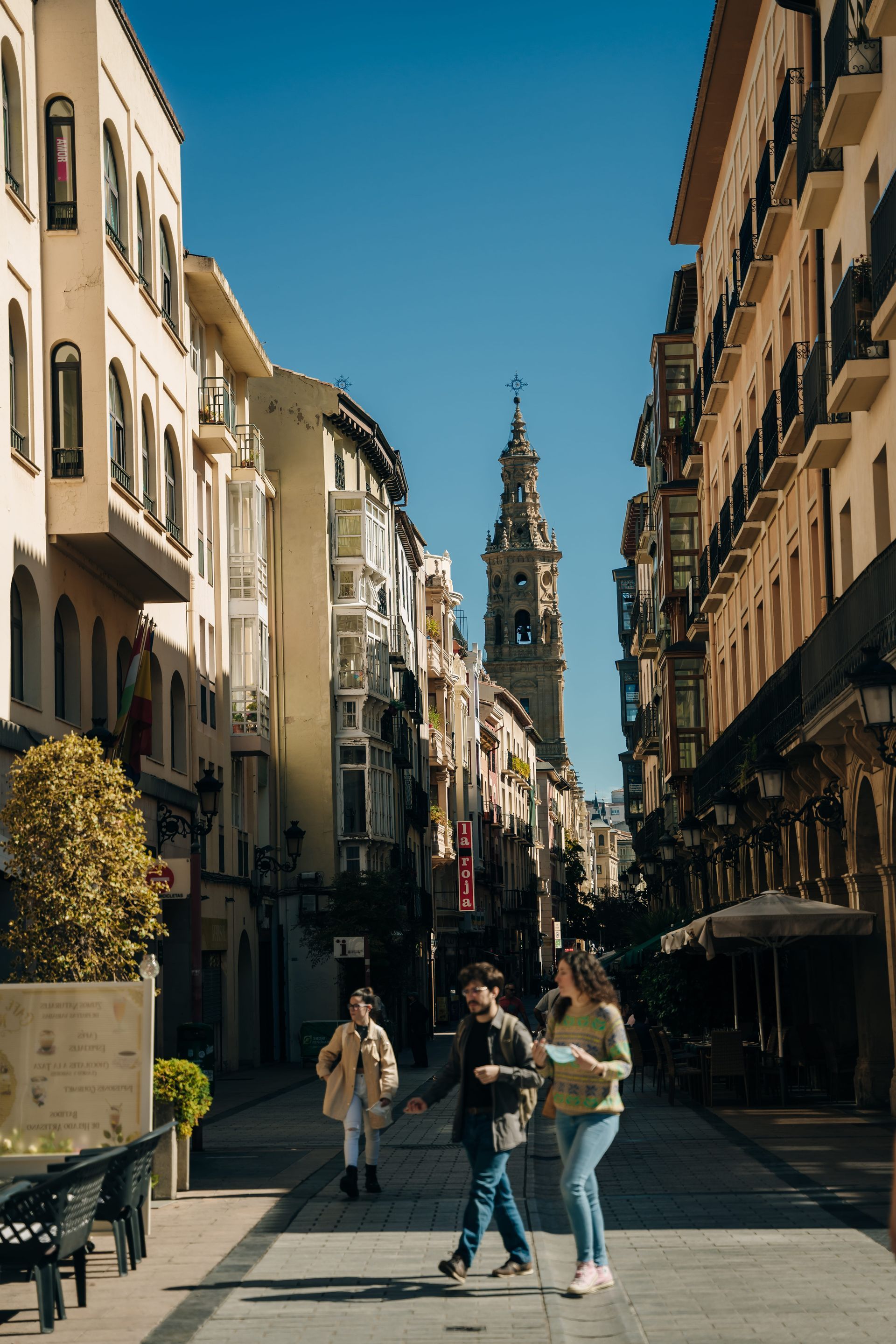 Tres personas caminan por una calle empedrada europea iluminada por el sol hacia la alta y ornamentada torre de una iglesia.