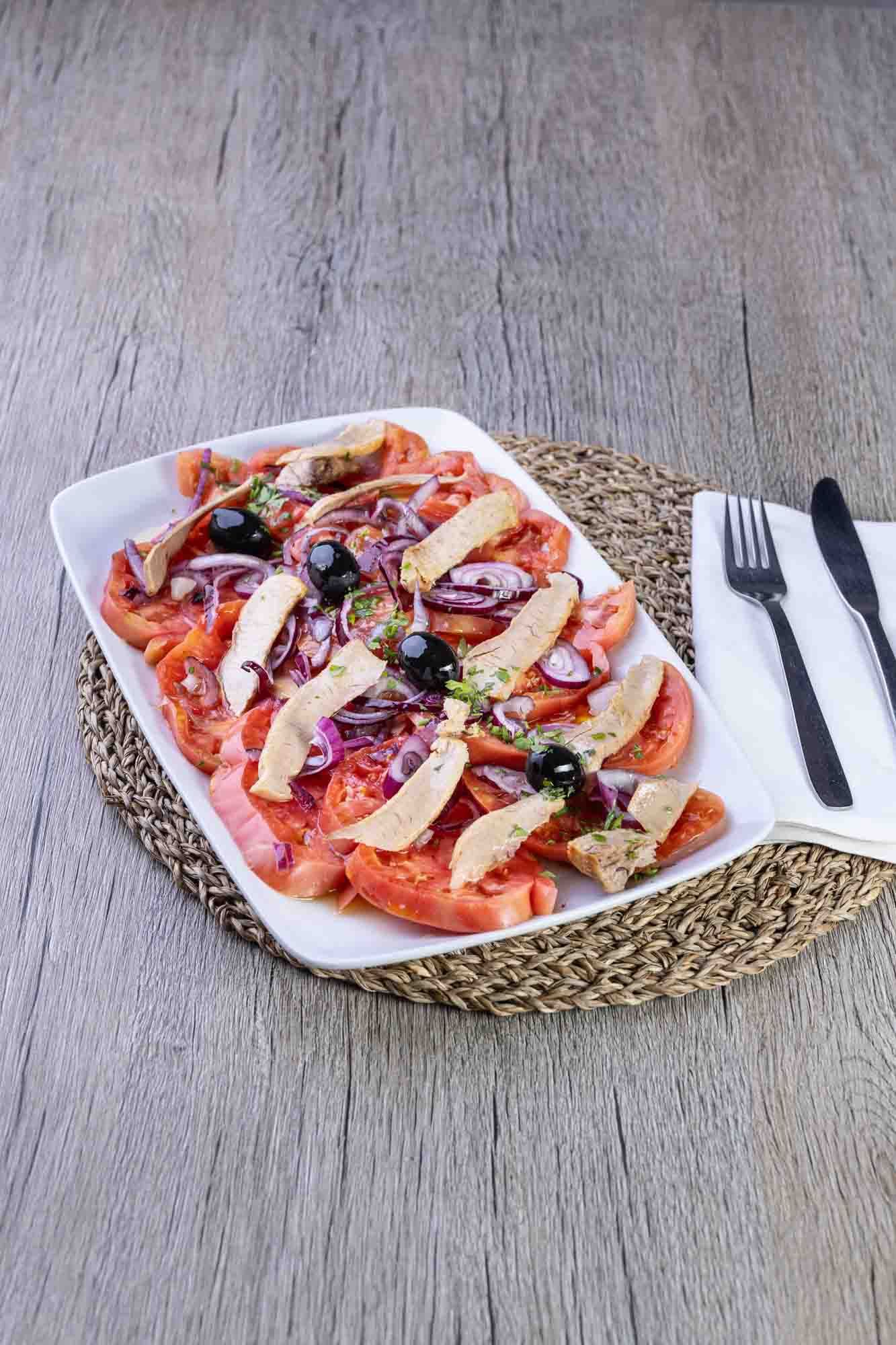 Un plato blanco cubierto con tomates, atún, aceitunas y cebollas.
