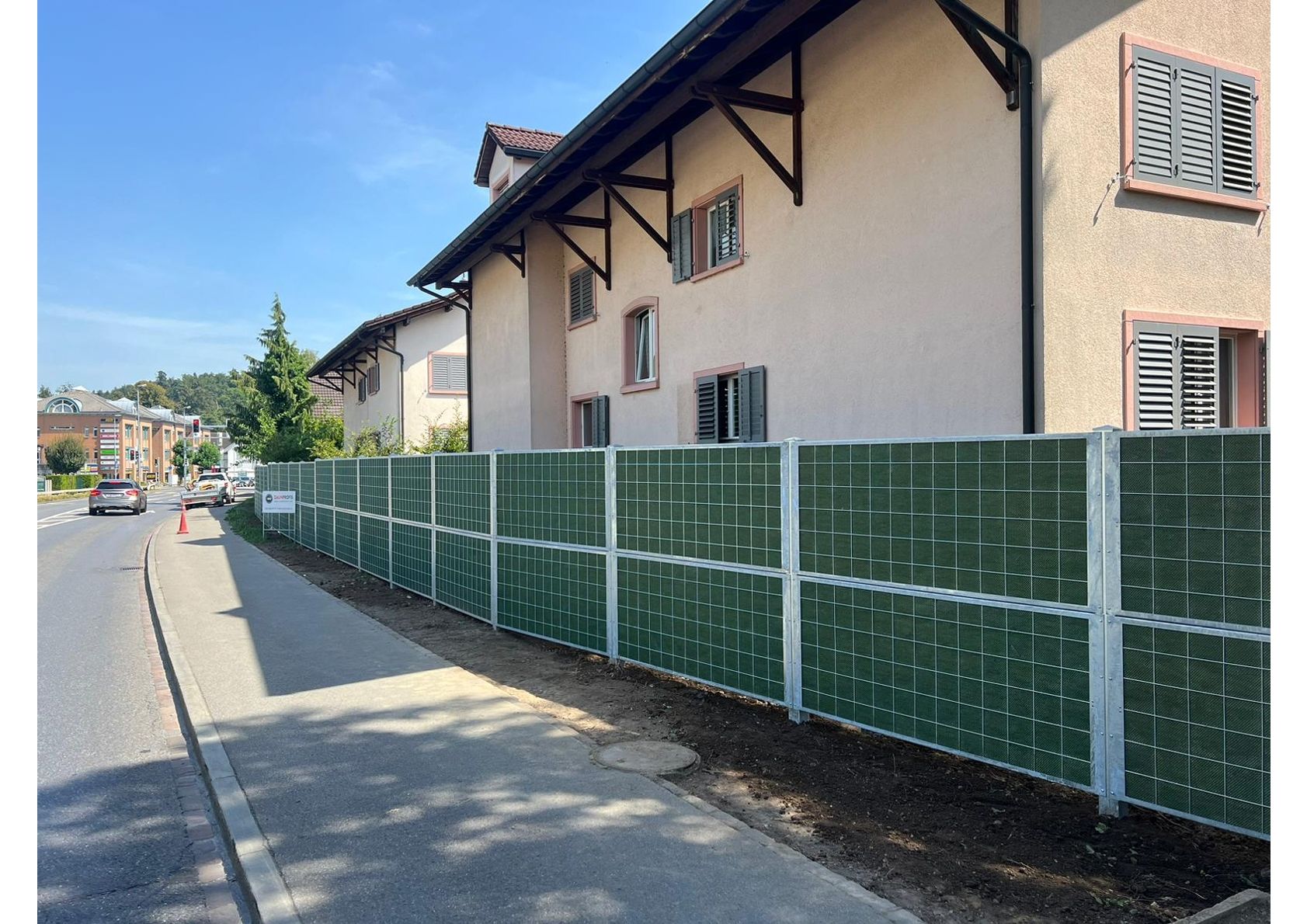 Grüne Lärmschutzwand entlang eines Gehwegs neben einer Straße, mit Häusern im Hintergrund unter einem sonnigen Himmel.