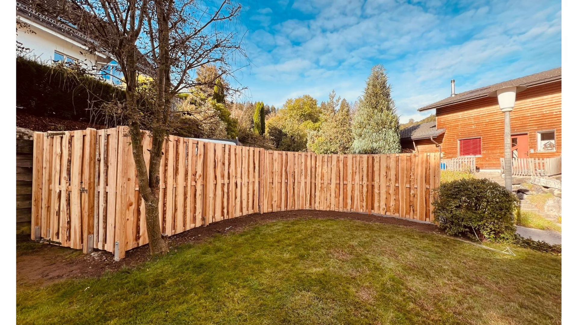 Ein Holzzaun, der sich über einen grasbewachsenen Hof schlängelt, mit einem Haus und Bäumen im Hintergrund.