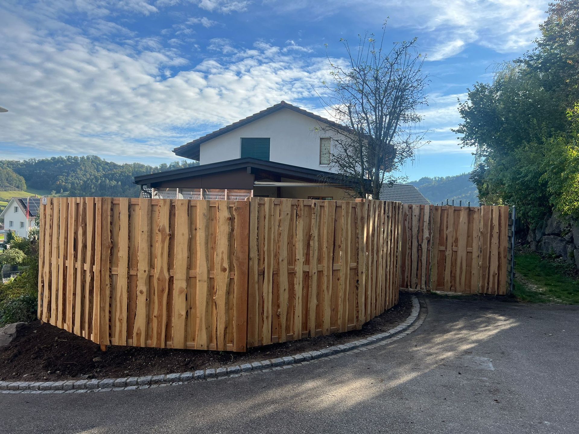 An einem sonnigen Tag schlängelt sich ein Holzzaun vor einem Haus entlang; Weg und Bäume sind sichtbar.
