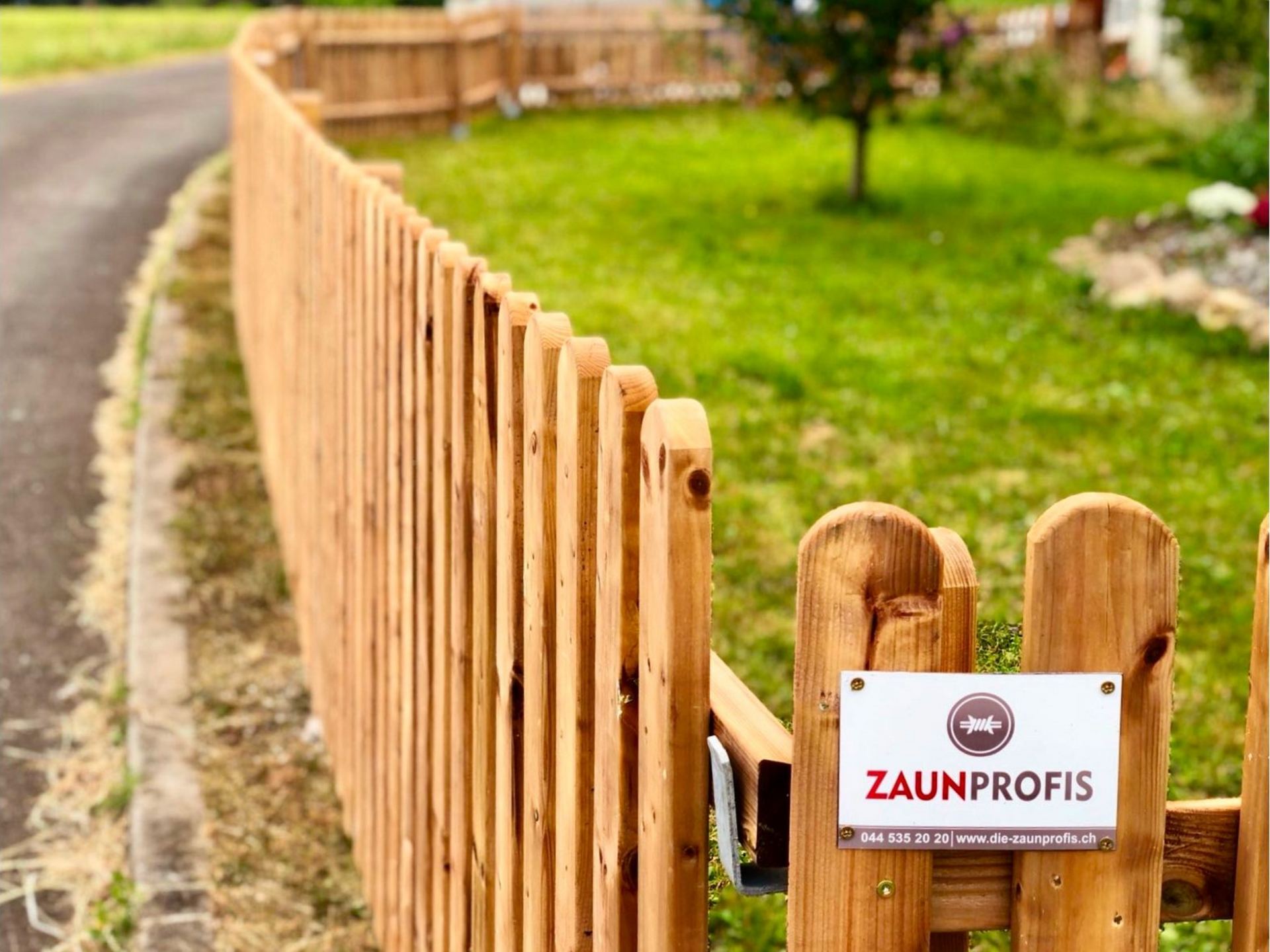 Holzzaun entlang einer Rasenfläche neben einer Straße. Schild „Zaunprofis“ am Tor.