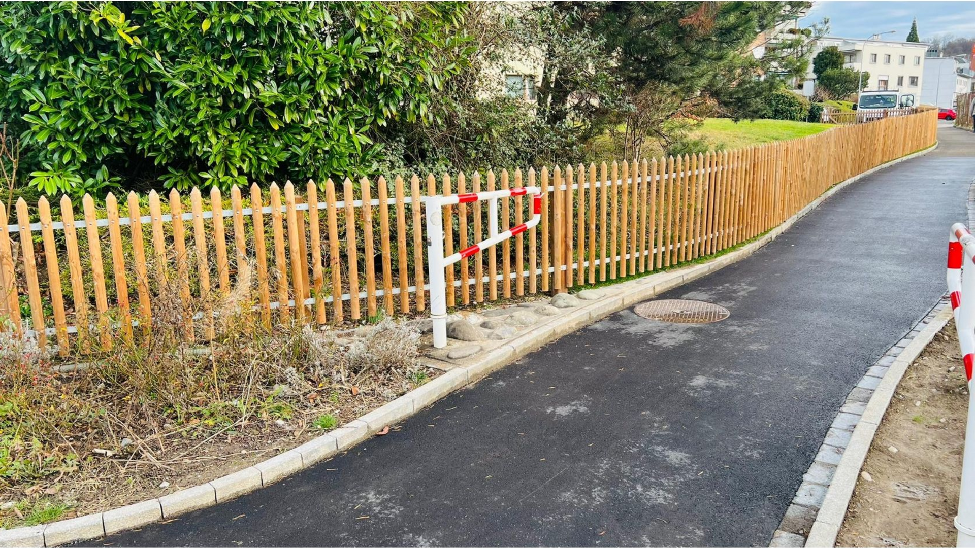 Ein gepflasterter Weg mit einem Holzzaun auf der linken Seite, einer Absperrung und einer Baumreihe im Hintergrund.