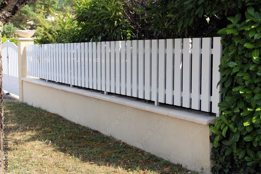 Weißer Lattenzaun auf einer beigen Mauer, mit einer grünen Hecke und Gras im Vordergrund.