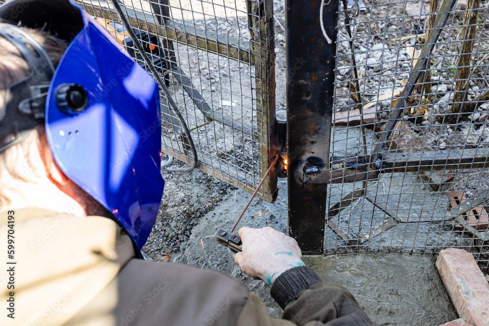 Person, die im Freien ein Metalltor schweißt und eine blaue Maske trägt.