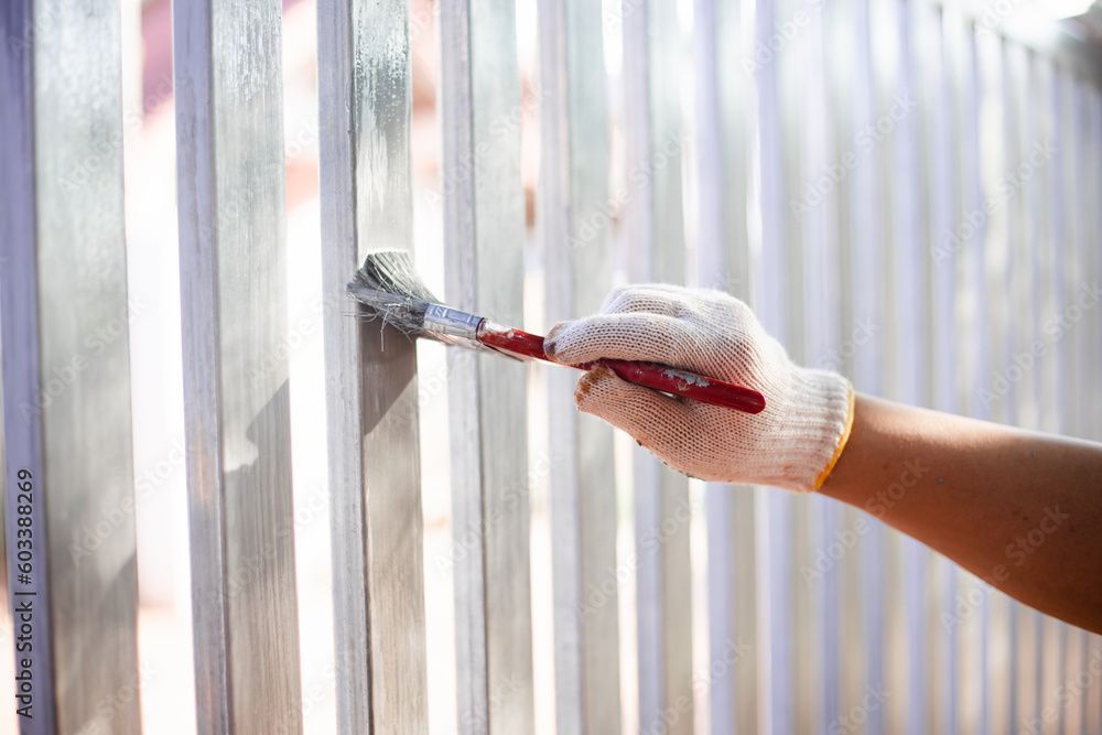 Person mit Handschuh, die mit einem Pinsel einen Metallzaun streicht.