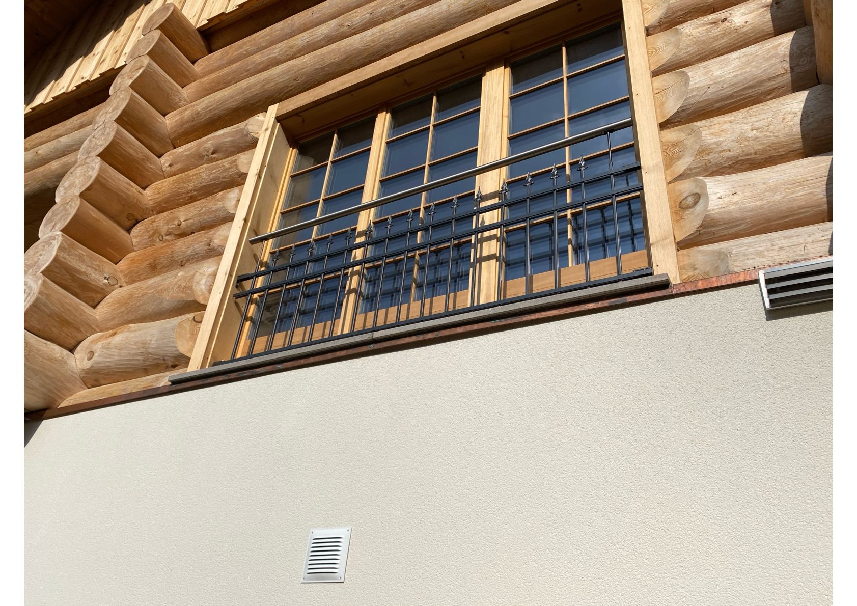Fenster mit schwarzen Gittern an einer Holzhütte vor einer strukturierten, hellen Wand.