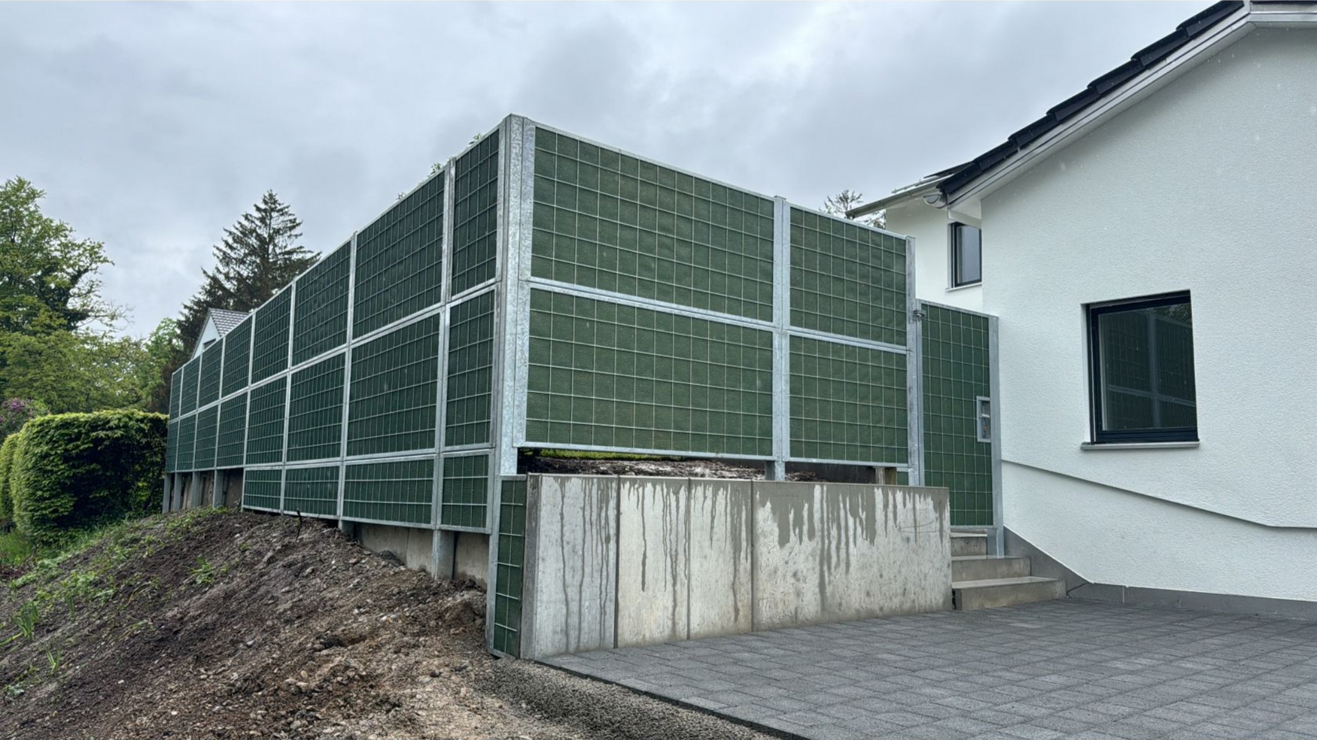 Grüne Schallschutzmauer entlang eines Betonfundaments neben einem weißen Haus an einem bewölkten Tag.