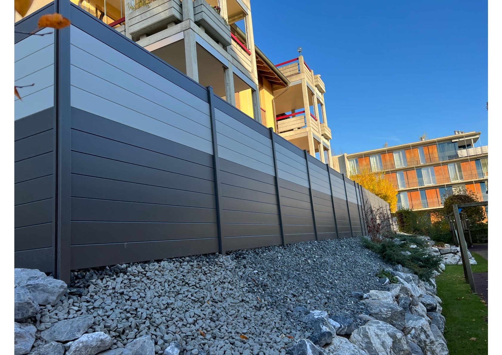 Grau-weißer Sichtschutzzaun aus Latten entlang eines Kies- und Felsdamms, im Hintergrund ein Gebäude.