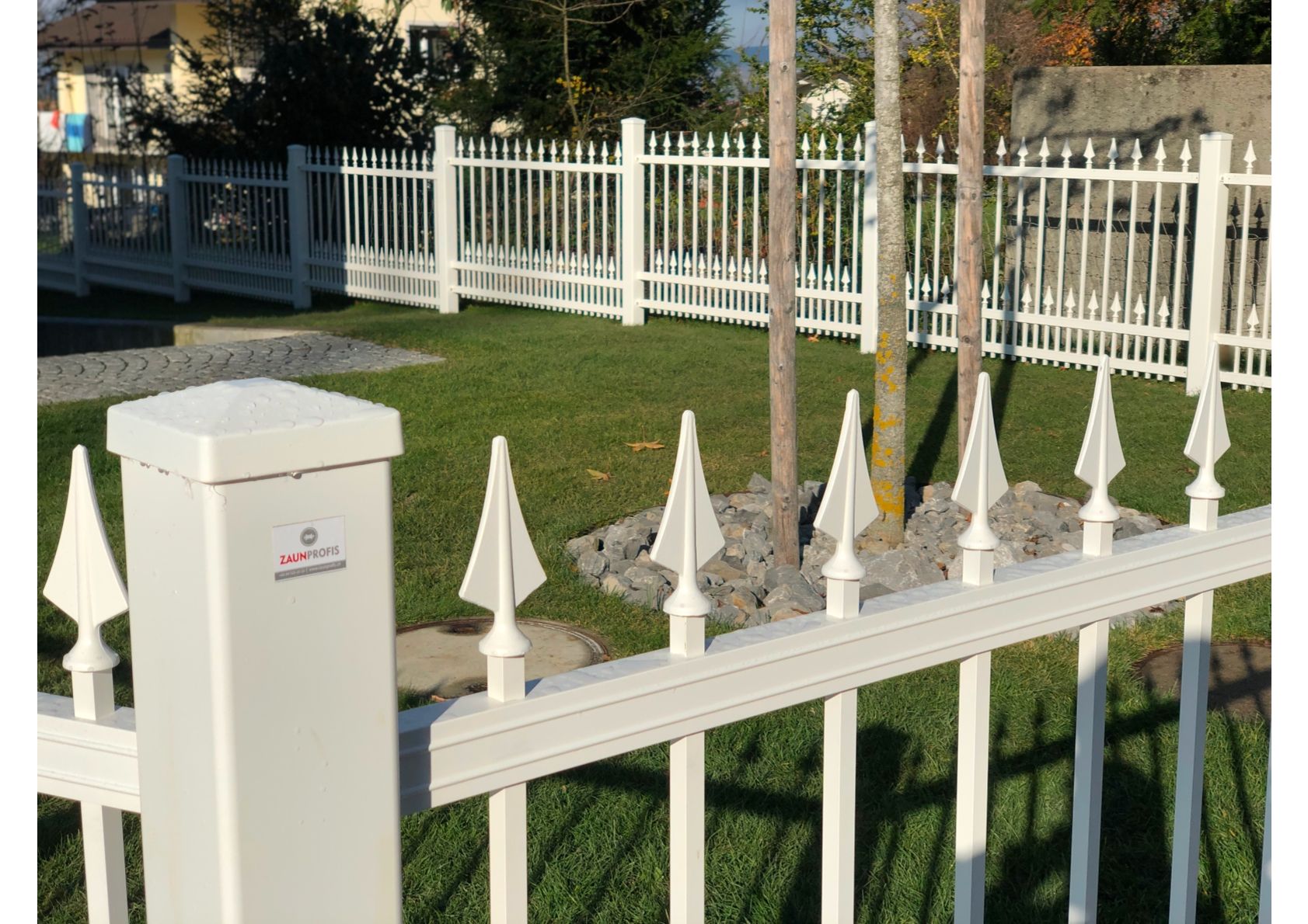 Weißer Metallzaun mit spitzen Spitzen, der einen grünen Rasen, Bäume und Gebäude im Hintergrund umgibt.