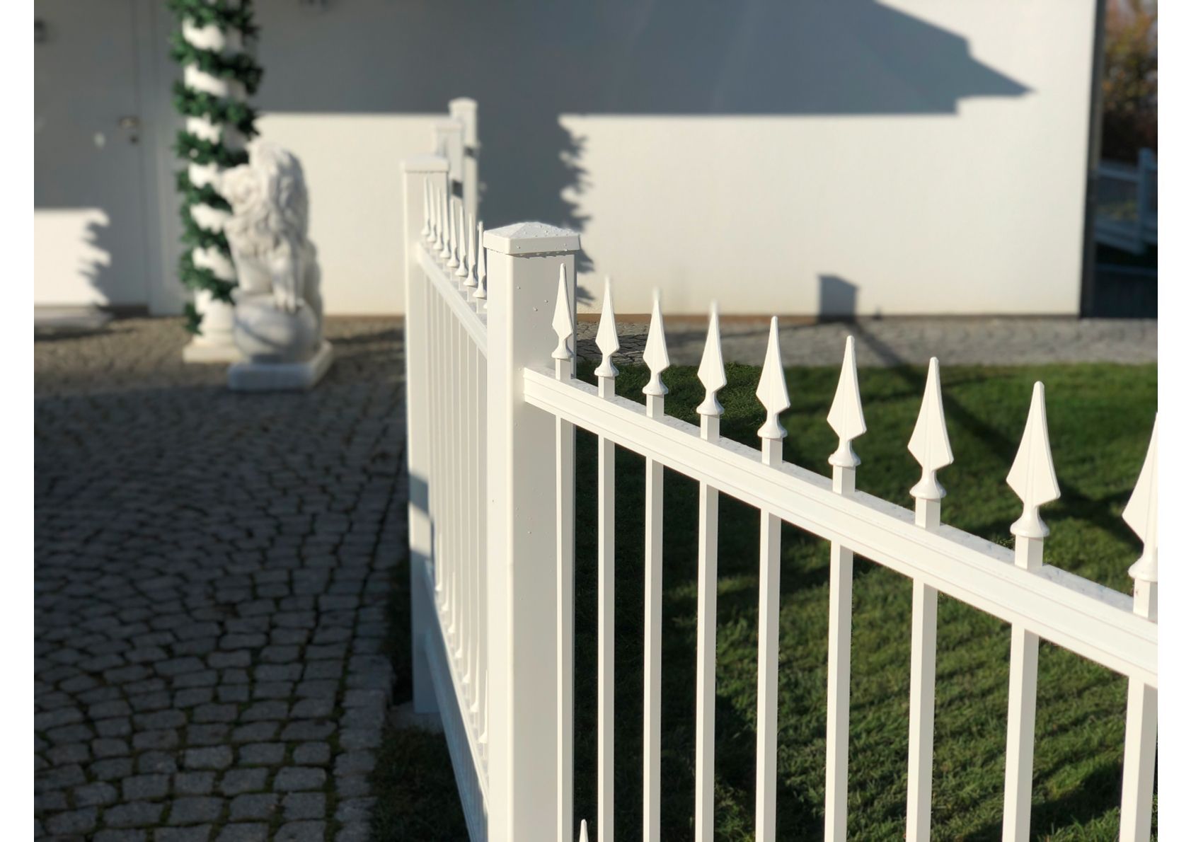 Ein weißer schmiedeeiserner Zaun mit speerartigen Endstücken grenzt an einen gepflasterten Gehweg und eine Rasenfläche.