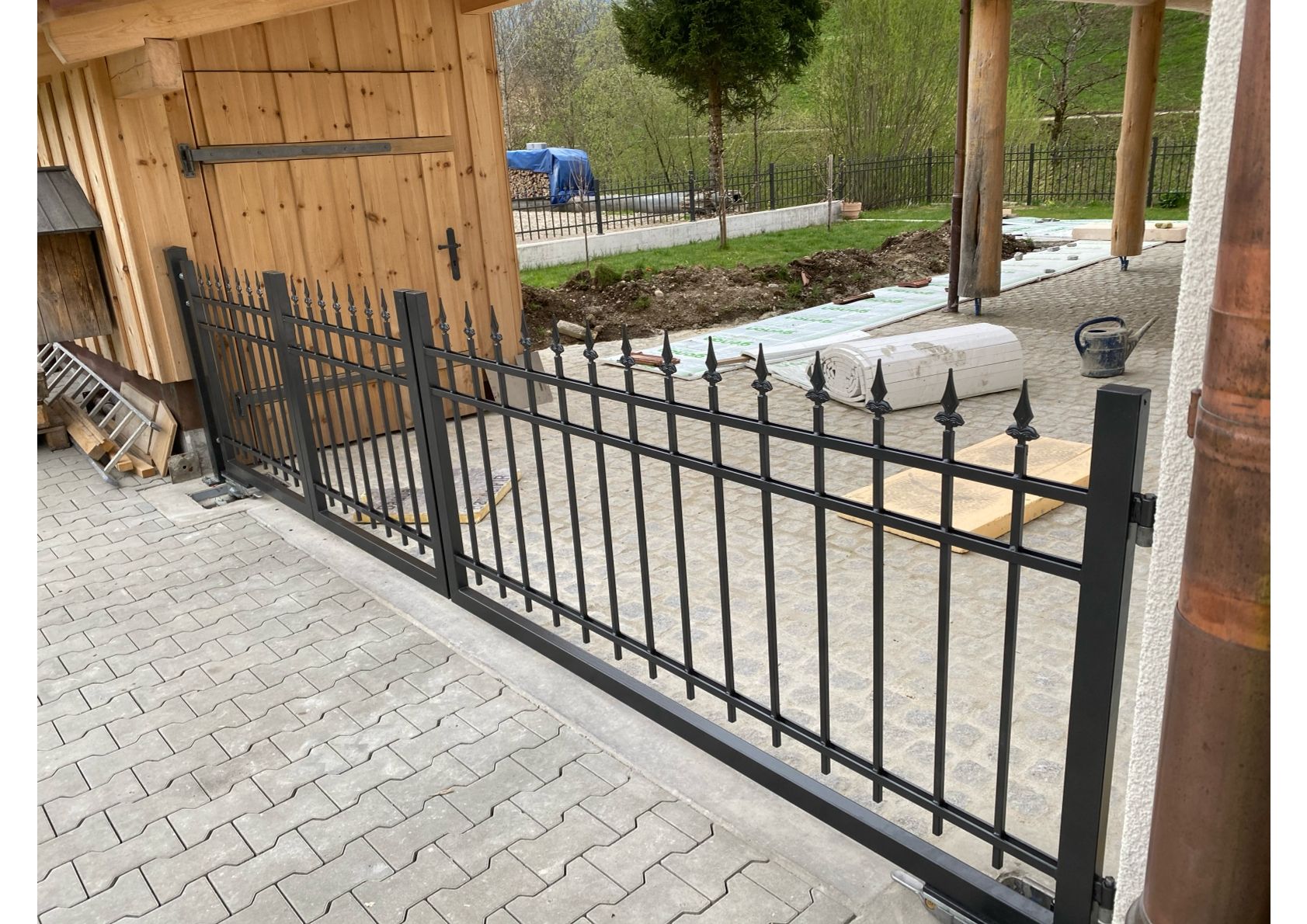 Schwarzer schmiedeeiserner Zaun vor einem Holzschuppen auf einer Steinterrasse.