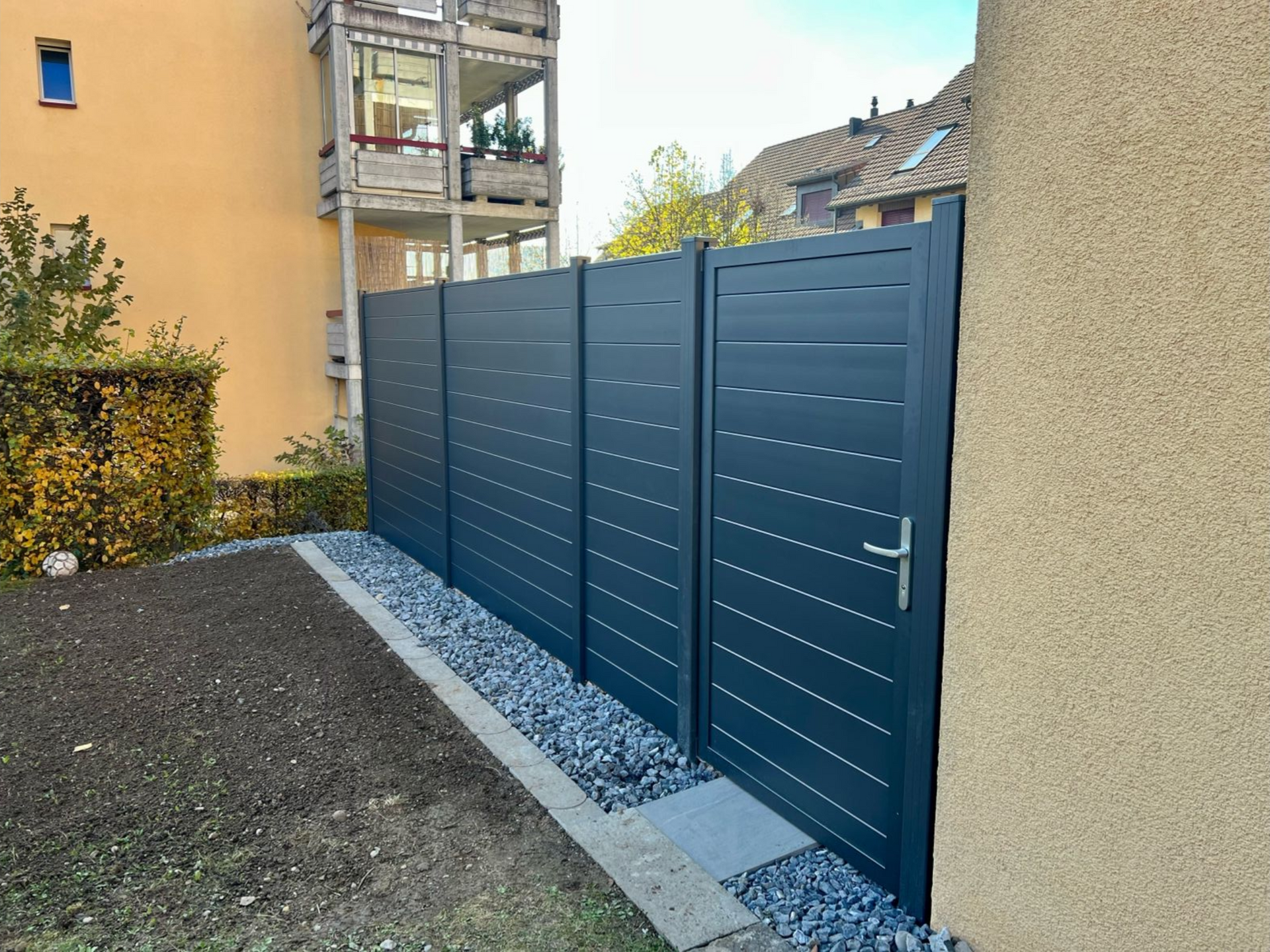 Dunkelgrauer Lattenzaun mit einer Tür, der an einen Hof mit Kies und einem beigen Gebäude grenzt.