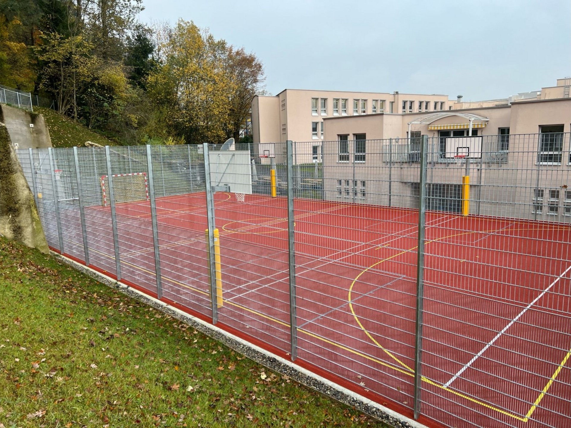 Roter Basketballplatz im Freien mit hohen Zäunen, neben einem Schulgebäude.