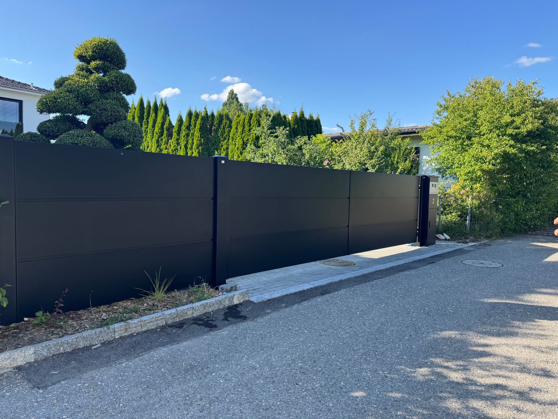 Schwarzer Metallzaun mit Tor an einer Schotterstraße, Bäume und blauer Himmel im Hintergrund.
