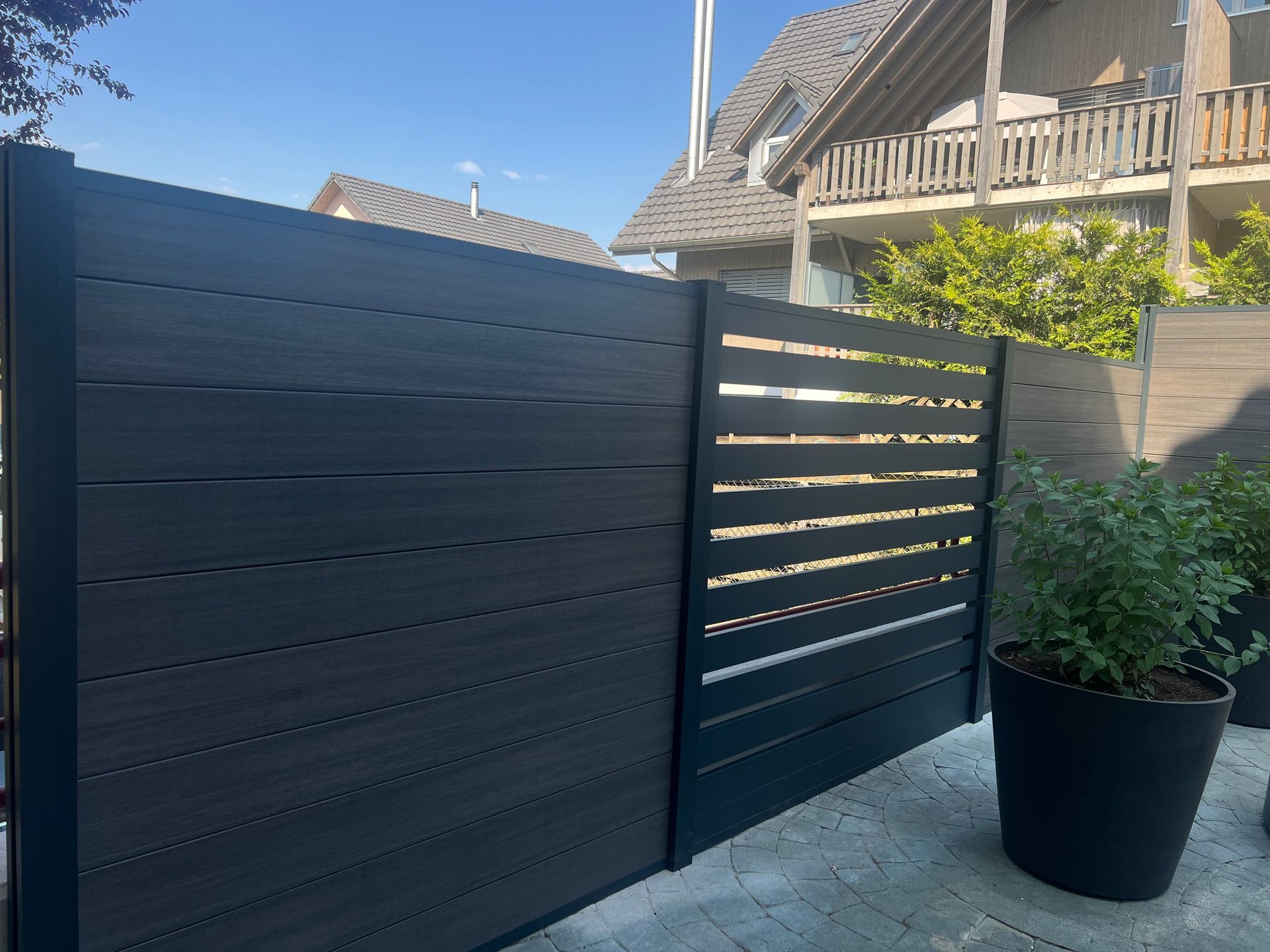 Dunkelgrauer Verbundzaun mit horizontalen, teilweise offenen Latten vor einem Haus und einer Topfpflanze.