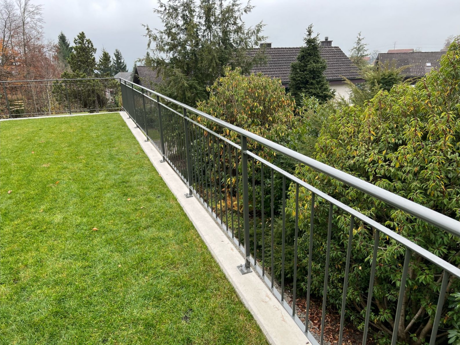 Rasen neben Metallgeländer und Büschen mit einem Wohnviertel im Hintergrund an einem bewölkten Tag.