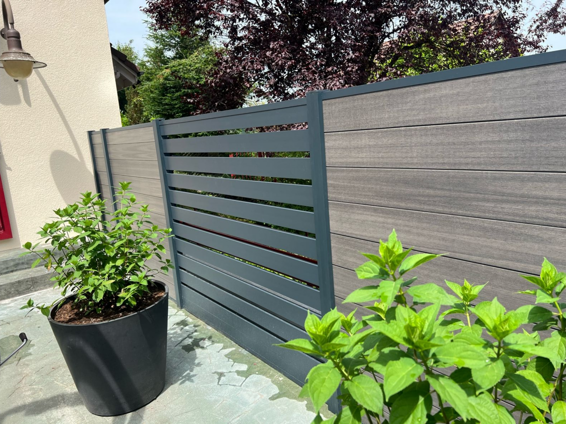 Verbundzaun mit grauen und holzgemaserten Paneelen, eingefasst von Pflanzen und einem dunklen Pflanzgefäß.