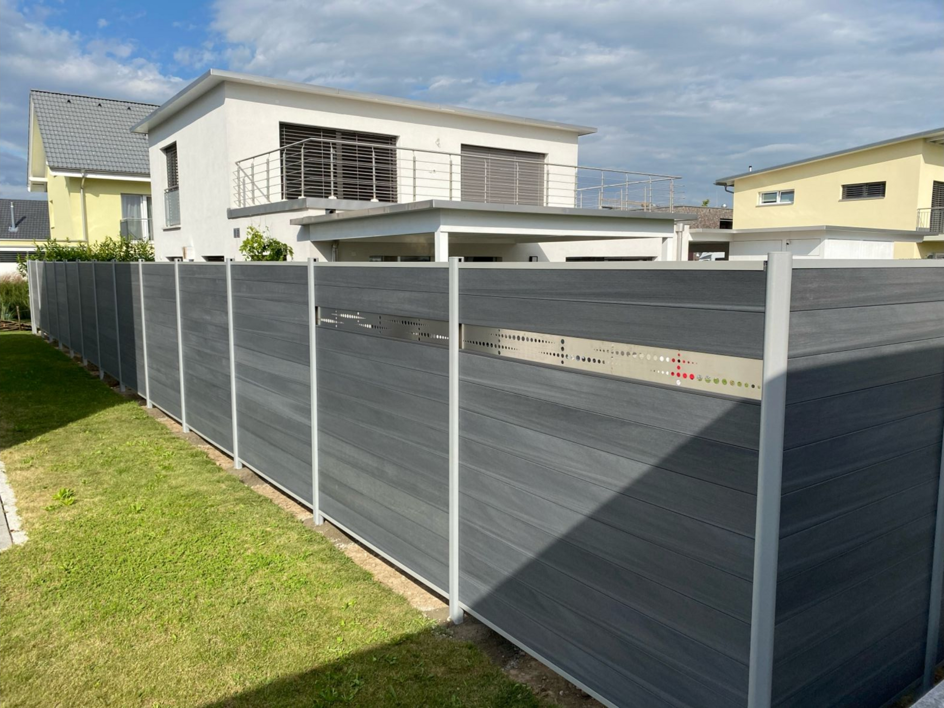 Grauer Sichtschutzzaun neben einem grasbewachsenen Hof mit Häusern im Hintergrund unter einem sonnigen Himmel.
