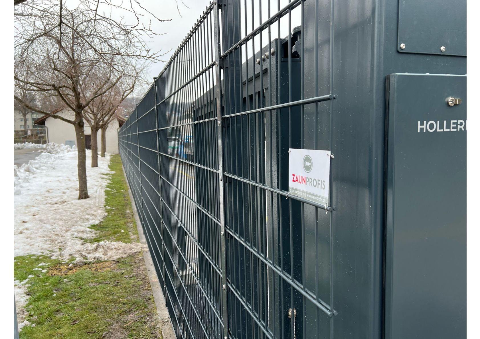 Schwarzer Metallzaun entlang einer Straße mit Bäumen und einem Gebäude im Hintergrund.