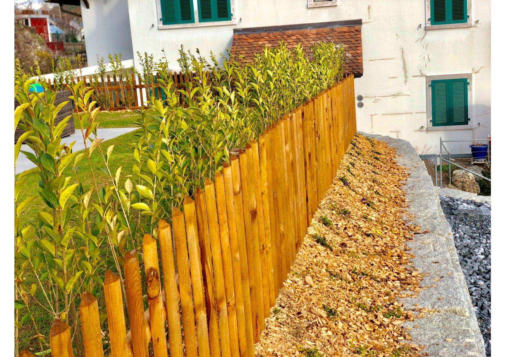 Holzzaun mit Grünpflanzen, neben einer mit Kies gesäumten Grenze, vor einem weißen Gebäude.