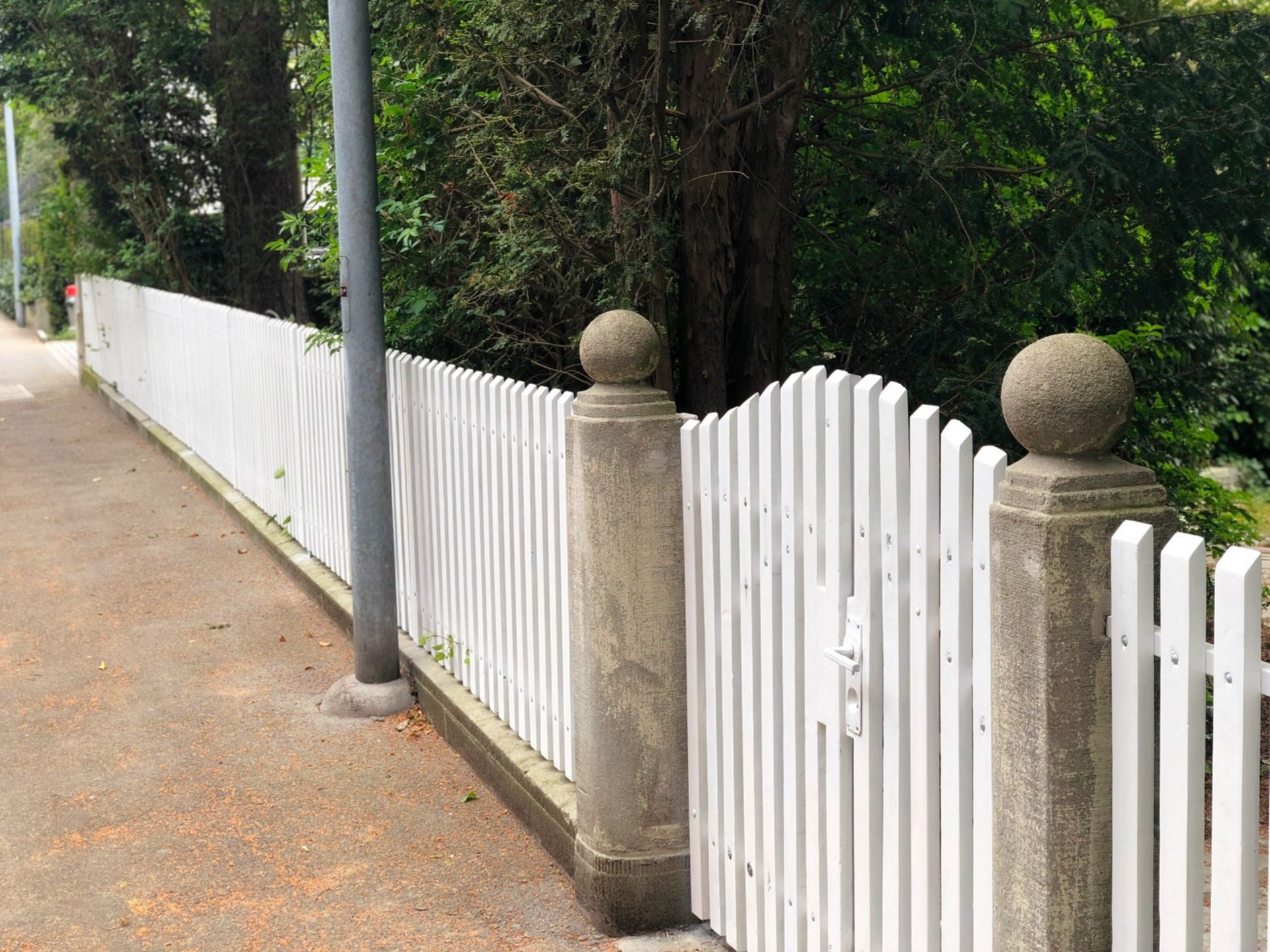 Weißer Lattenzaun mit Steinsäulen, der an einen Gehweg und Grünflächen grenzt.