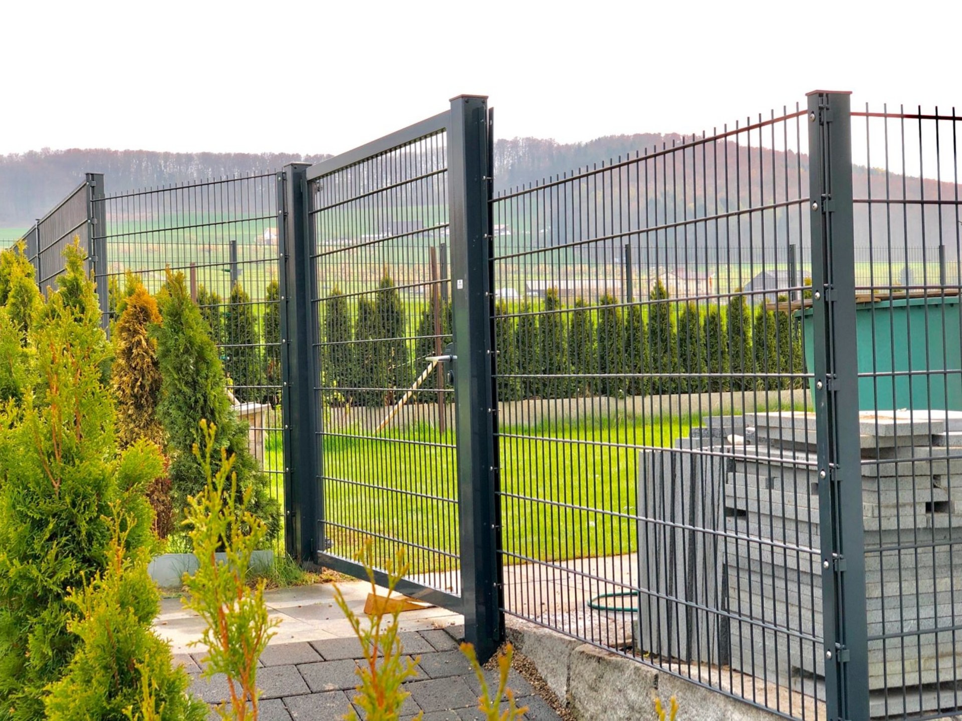 Grauer Metallzaun mit Tor, davor Gebüsch und Steinweg, im Hintergrund ein grasbewachsener Hof und eine Baumreihe.