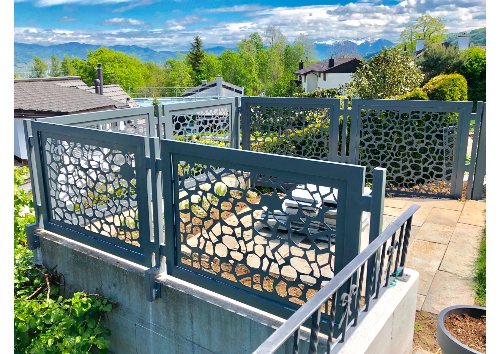 Grauer dekorativer Metallzaun mit kieselsteinartigen Aussparungen und Blick auf die malerische Berglandschaft.