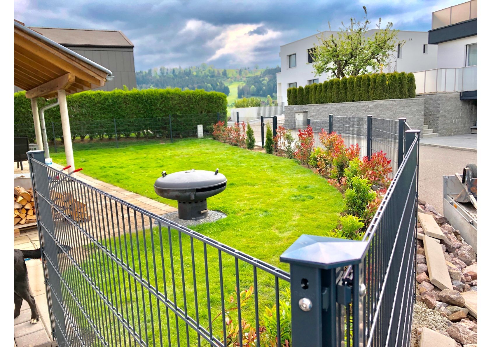 Eine eingezäunte grüne Wiese mit Grill und kleinen Bäumen, die an ein Haus in einem sonnigen Wohngebiet grenzt.