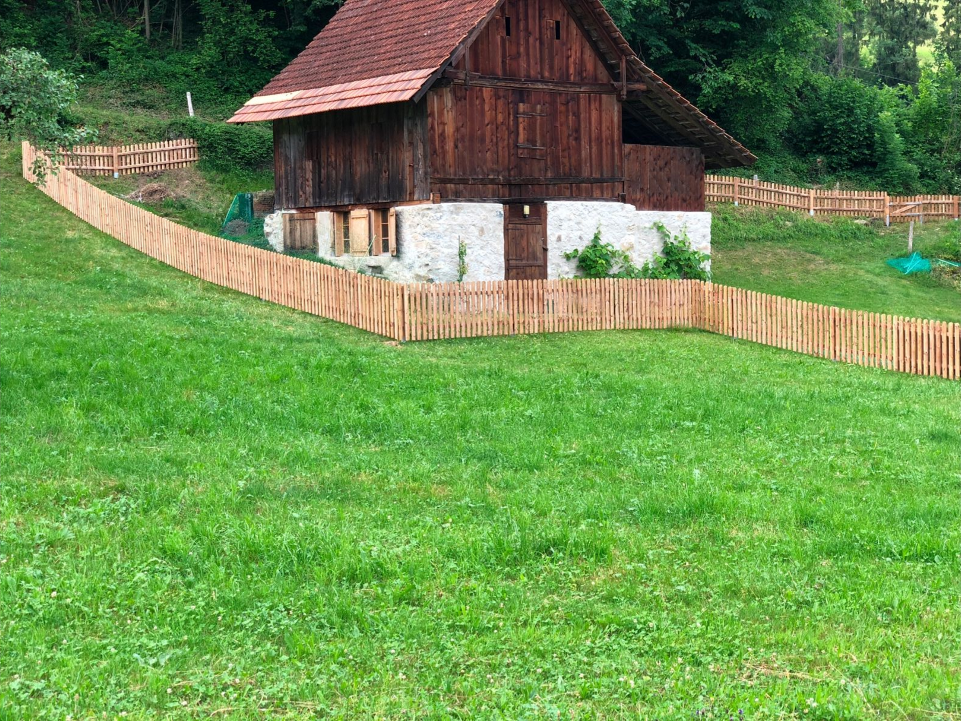Holzzaun in einem grasbewachsenen Hof, dahinter eine braune Scheune, im Hintergrund grüne Bäume.
