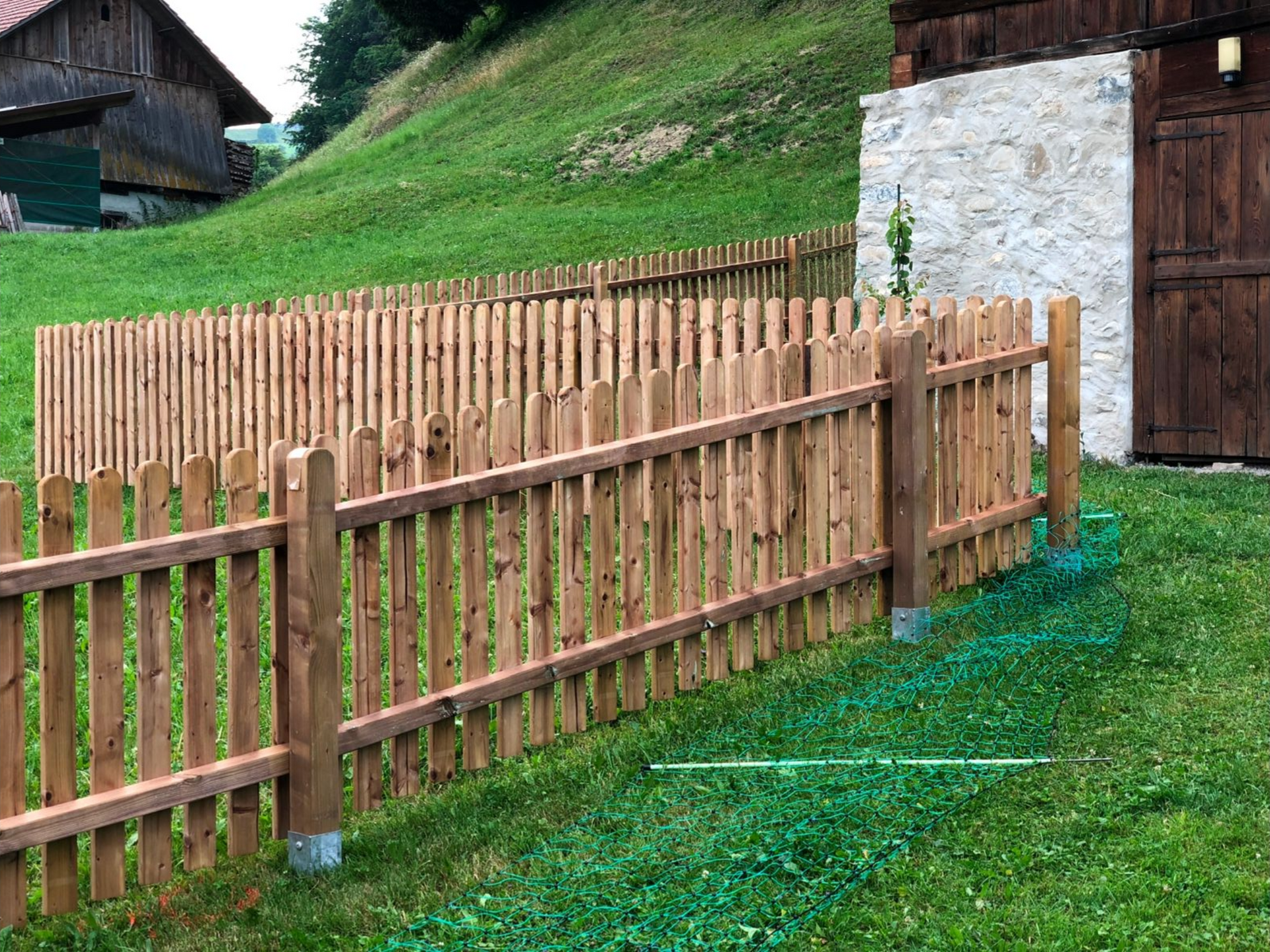 Holzlattenzaun in einem grasbewachsenen Hof, in der Nähe einer braunen Scheune und eines grünen Hügels.