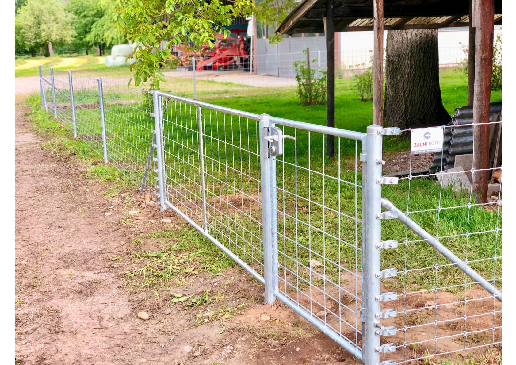 Metallzaun mit Tor, der eine Rasenfläche neben einem Feldweg schützt. Roter Traktor im Hintergrund.