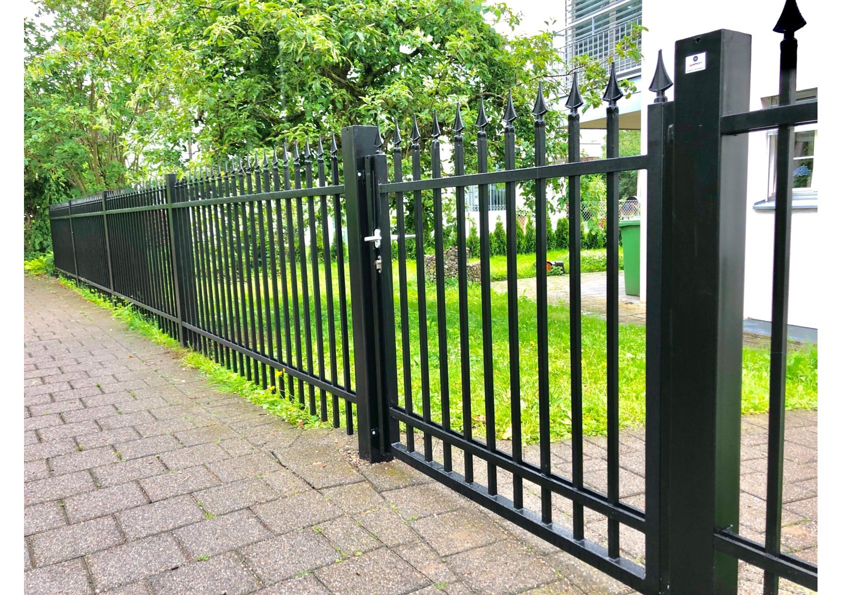 Schwarzer Metallzaun mit einem Tor auf einem gemauerten Gehweg vor grünem Gras und Bäumen.