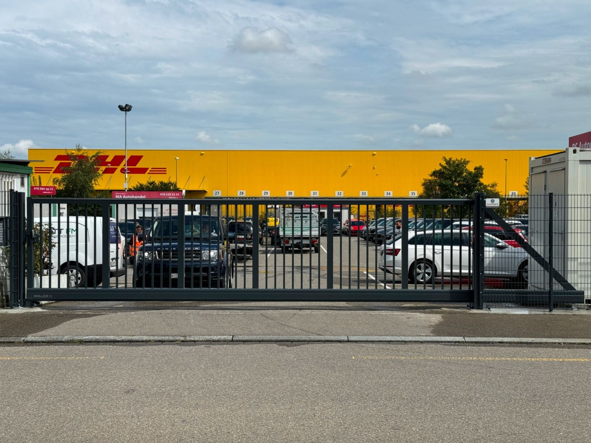 Schwarzes Tor vor gelbem DHL-Gebäude mit parkenden Autos. Bewölkter Himmel.