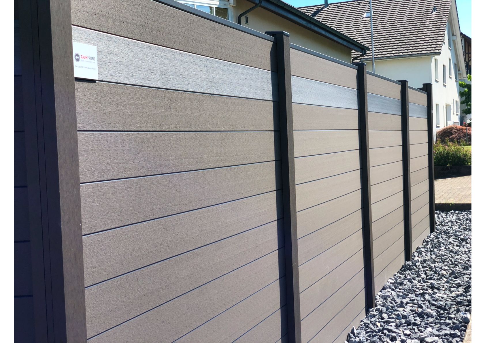 Braun-grauer Verbundzaun mit silbernem Streifen entlang eines Hofes mit Kies.