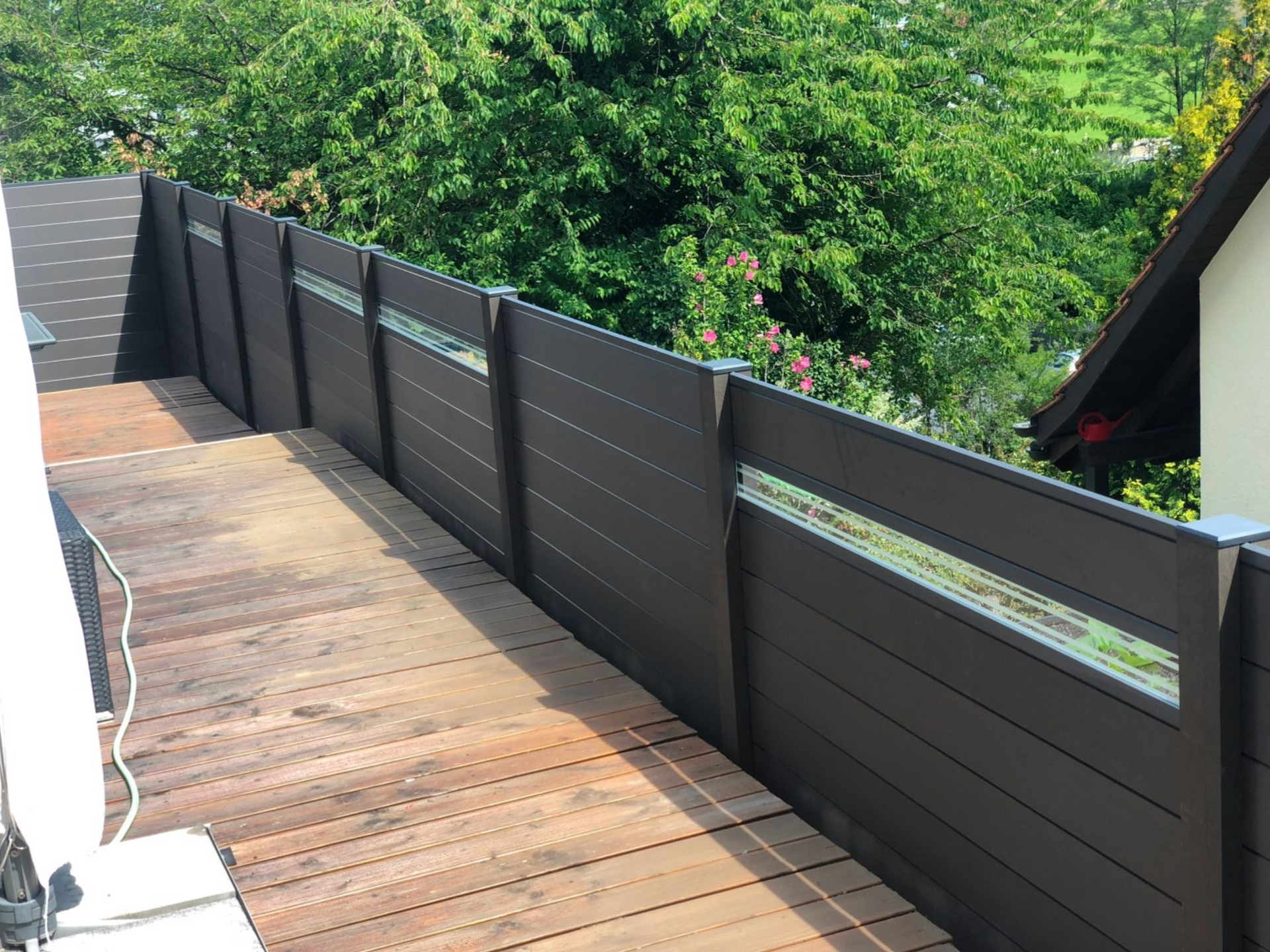 Brauner Verbundzaun auf Holzterrasse; Bäume im Hintergrund.